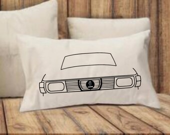 May include: Cream-coloured rectangular cushion featuring a black line drawing of a car's front end. The design includes the bonnet, grille, headlights, and a logo. The cushion is resting on a wooden surface.