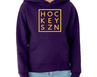May include: A purple hoodie with a front pocket and a hood. The hoodie displays the text "HOCKEY SZN" in a yellow square. The person in the image is wearing blue jeans.