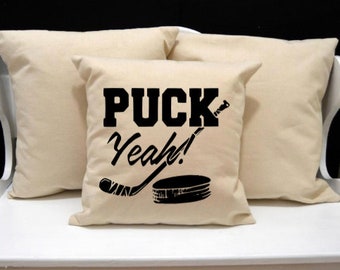 May include: Three cream-coloured throw cushions. The central cushion features the words "PUCK Yeah!" in black, with a hockey stick and puck graphic. The cushions are arranged on a white surface against a black backdrop.