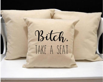 May include: Three beige square cushions. The central cushion features the phrase "Bitch, TAKE A SEAT" in black script. The cushions are made of a soft fabric and are arranged on a white surface.