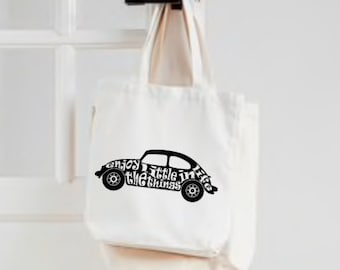 May include: A cream-coloured tote bag featuring a black graphic of a classic car. The car graphic incorporates the phrase "enjoy the little things" in a decorative font. The bag is suspended on a white door.