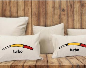 May include: Several cream-coloured throw cushions of various sizes are arranged on a wooden surface. Two cushions have a graphic of a curved gauge with yellow and red sections, and the word "turbo" below. The background is a wooden wall.
