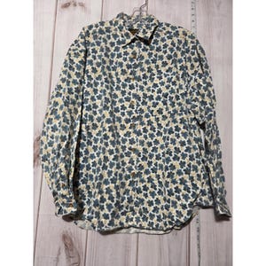 Pode incluir: Uma camisa de manga comprida com botões e um padrão de folhas em tons de azul, bege e branco. A camisa tem uma gola clássica e botões de madeira. A camisa está pendurada num cabide.