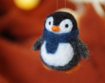 May include: A handmade felt penguin ornament. The penguin has a black head and wings, a white belly, and an orange beak and feet. It wears a blue scarf. The background is a blurred orange.