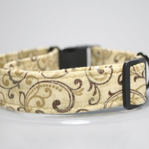 Puede incluir: Un collar para perros beige con un patrón repetido de espirales marrones y doradas. El collar tiene una hebilla de plástico negro y una anilla en D de metal negro para sujetar la correa. La tela parece ser de algodón suave.