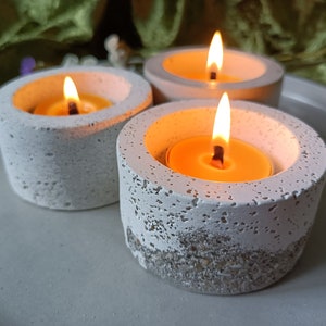 Puede incluir: Tres velas de té encendidas en portavelas de hormigón gris. Los portavelas tienen un aspecto texturizado y moteado con una base de color más claro. Las velas están sobre un plato gris claro, con un fondo verde.