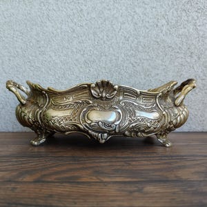 May include: An ornate silver-toned metal centerpiece bowl with decorative handles and feet. The bowl features intricate embossed patterns and a scalloped rim. A classic decorative piece for the home.