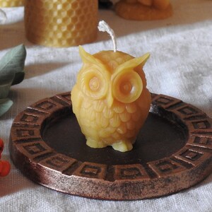 May include: A small, yellow beeswax candle shaped like an owl. The candle is sitting on a brown, circular, decorative plate with a raised edge.