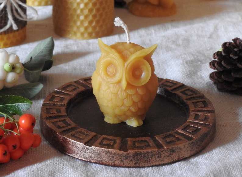 May include: A small, yellow beeswax candle shaped like an owl. The candle is sitting on a brown, circular, decorative plate with a raised edge.