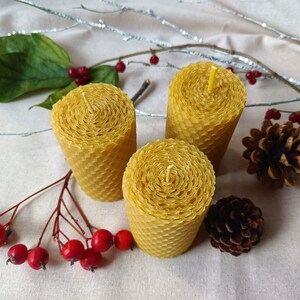 Puede incluir: Tres velas de panal de abeja amarillas sobre un fondo de tela blanca con bayas rojas y pi&ntilde;as.