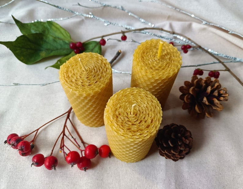 Puede incluir: Tres velas de panal de abeja amarillas sobre un fondo de tela blanca con bayas rojas y pi&ntilde;as.