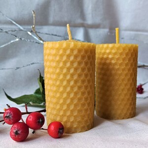 Puede incluir: Dos velas amarillas de cera de abeja con un patr&oacute;n de panal. Las velas est&aacute;n sobre una superficie blanca con bayas rojas y hojas verdes.