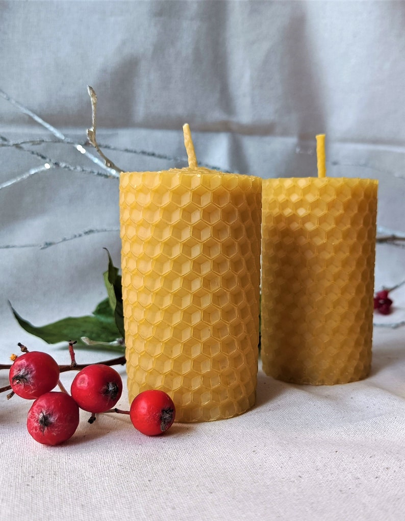 Puede incluir: Dos velas amarillas de cera de abeja con un patr&oacute;n de panal. Las velas est&aacute;n sobre una superficie blanca con bayas rojas y hojas verdes.