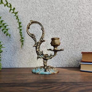 Puede incluir: Candelabro de latón ornamentado con base floral y asa decorativa. El candelabro tiene un aspecto envejecido con detalles turquesa en la base. El diseño es detallado e intrincado, con una altura aproximada de 15 cm.