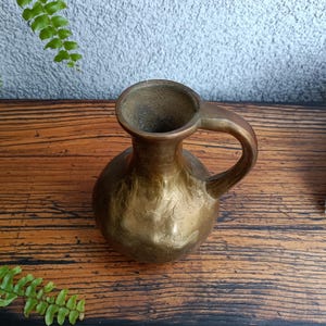 Op de afbeelding: Een kleine, gouden messing vaas met een getextureerd oppervlak en een enkele handgreep. De vaas staat op een houten oppervlak.