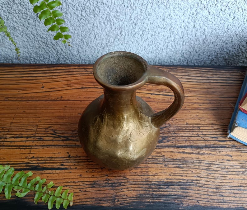 Op de afbeelding: Een kleine, gouden messing vaas met een getextureerd oppervlak en een enkele handgreep. De vaas staat op een houten oppervlak.