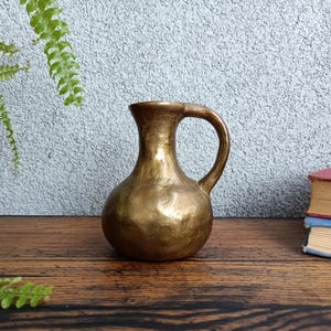 Op de afbeelding: Een kleine, gouden metalen kan met een gehamerde textuur en een rond lichaam. De kan heeft &eacute;&eacute;n handvat en een smalle opening.