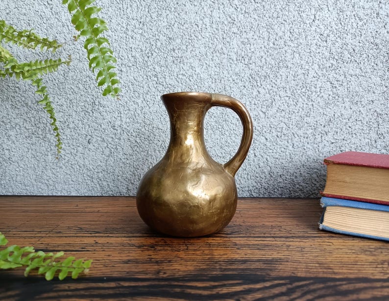 Op de afbeelding: Een kleine, gouden metalen kan met een gehamerde textuur en een rond lichaam. De kan heeft &eacute;&eacute;n handvat en een smalle opening.