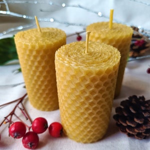 Puede incluir: Tres velas de cera de abeja amarilla con un patr&oacute;n de panal y mechas naturales. Las velas est&aacute;n dispuestas sobre una superficie blanca con bayas rojas y una pi&ntilde;a.