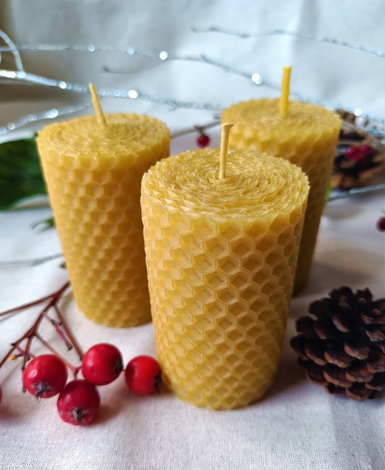 Puede incluir: Tres velas de cera de abeja amarilla con un patr&oacute;n de panal y mechas naturales. Las velas est&aacute;n dispuestas sobre una superficie blanca con bayas rojas y una pi&ntilde;a.