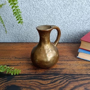 Op de afbeelding: Een gouden keramische vaas met een getextureerd oppervlak en een handvat. De vaas staat op een houten tafel.