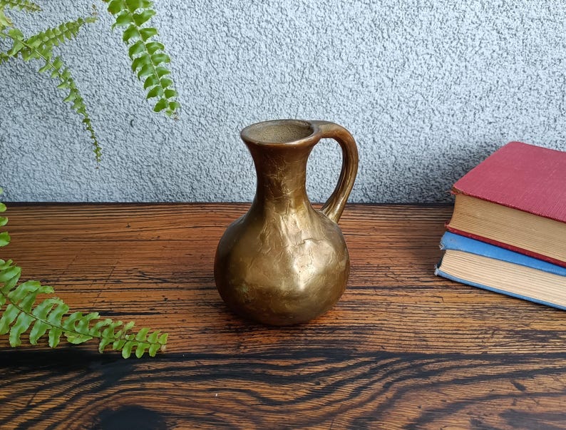 Op de afbeelding: Een gouden keramische vaas met een getextureerd oppervlak en een handvat. De vaas staat op een houten tafel.