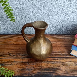Op de afbeelding: Een kleine, gouden metalen kan met een handvat. De kan heeft een getextureerd oppervlak en staat op een houten oppervlak.