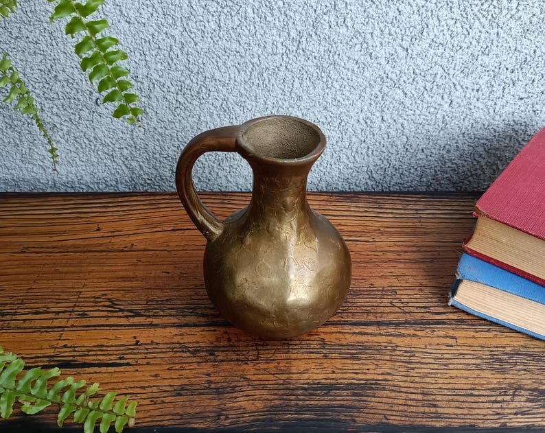 Op de afbeelding: Een kleine, gouden metalen kan met een handvat. De kan heeft een getextureerd oppervlak en staat op een houten oppervlak.