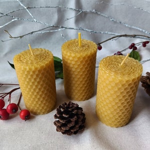 Puede incluir: Tres velas de cera de abeja amarilla con un patr&oacute;n de panal. Las velas est&aacute;n sobre una superficie de tela blanca con una peque&ntilde;a pi&ntilde;a y bayas rojas.