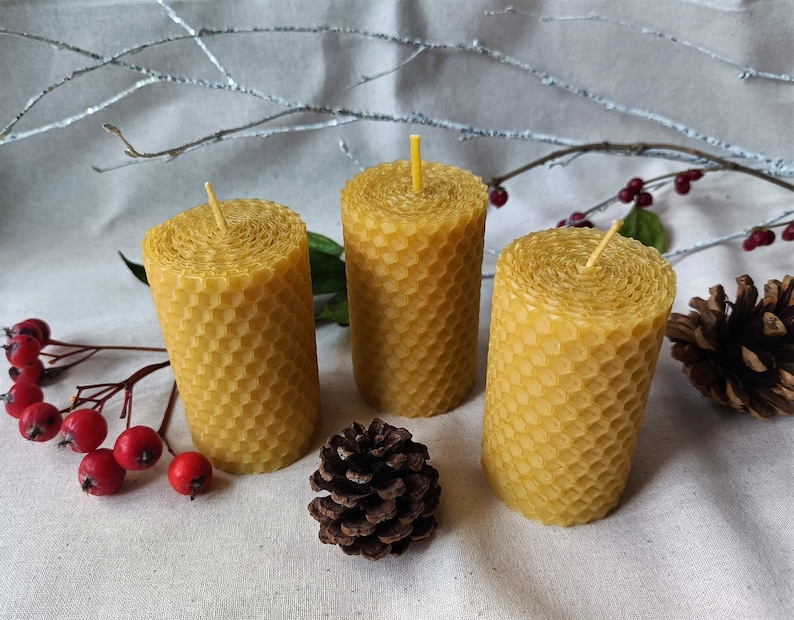 Puede incluir: Tres velas de cera de abeja amarilla con un patr&oacute;n de panal. Las velas est&aacute;n sobre una superficie de tela blanca con una peque&ntilde;a pi&ntilde;a y bayas rojas.