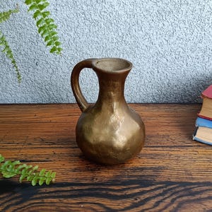 Op de afbeelding: Een kleine, gouden metalen kan met een gehamerde afwerking. De kan heeft een rond lichaam en een gebogen handvat.