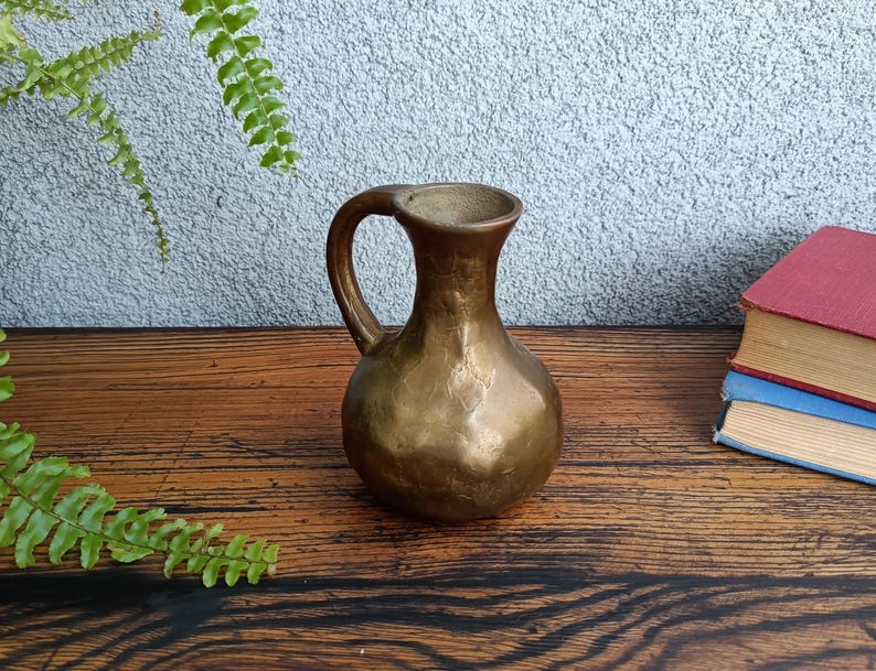 Op de afbeelding: Een kleine, gouden metalen kan met een gehamerde afwerking. De kan heeft een rond lichaam en een gebogen handvat.