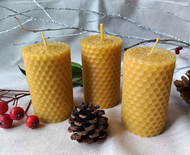 Puede incluir: Tres velas de cera de abeja amarilla con un patr&oacute;n de panal. Las velas est&aacute;n sobre una superficie blanca con una peque&ntilde;a pi&ntilde;a delante de ellas.
