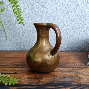Op de afbeelding: Een kleine, gouden messing kan met een rond lichaam en een enkele handgreep. De kan heeft een getextureerd oppervlak en staat op een houten tafel.