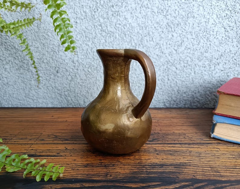 Op de afbeelding: Een kleine, gouden messing kan met een rond lichaam en een enkele handgreep. De kan heeft een getextureerd oppervlak en staat op een houten tafel.