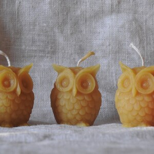 May include: Three small, yellow beeswax candles shaped like owls. The candles have a white wick and are sitting on a white linen fabric.