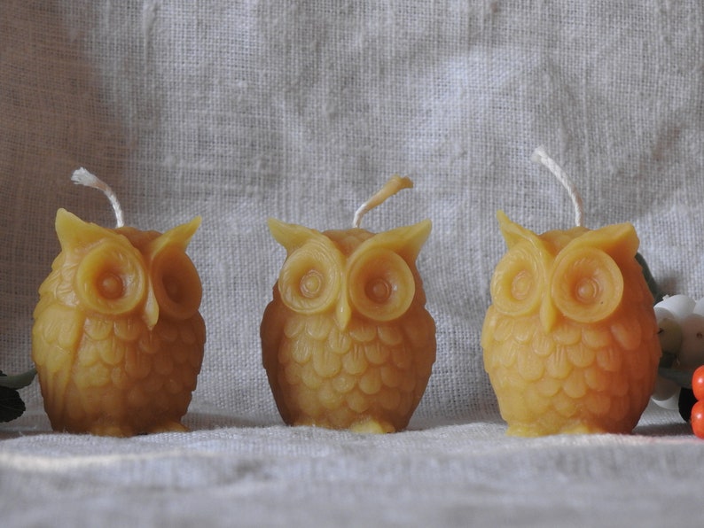 May include: Three small, yellow beeswax candles shaped like owls. The candles have a white wick and are sitting on a white linen fabric.