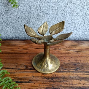 Puede incluir: Un candelero de latón con diseño de hojas. El candelero tiene una base redonda y un tallo central que sostiene seis hojas estilizadas. Las hojas están dispuestas en un patrón circular, y el diseño general recuerda a una flor.