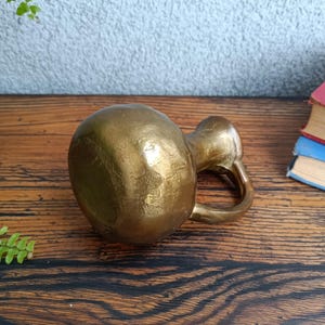 Op de afbeelding: Een kleine, gouden metalen vaas met een getextureerd oppervlak. De vaas heeft een rond lichaam en een korte, gebogen tuit.