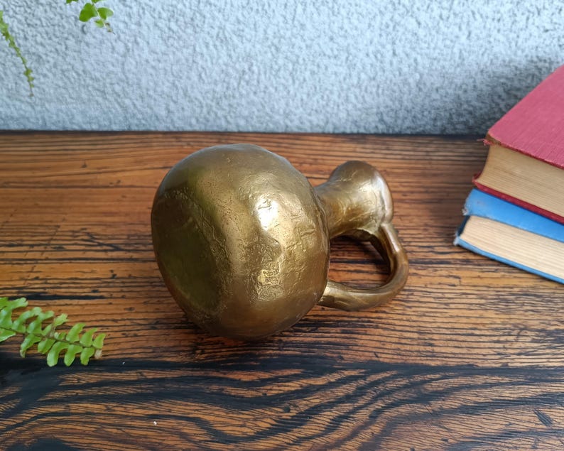 Op de afbeelding: Een kleine, gouden metalen vaas met een getextureerd oppervlak. De vaas heeft een rond lichaam en een korte, gebogen tuit.