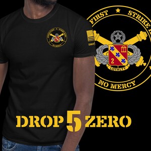 May include: Black t-shirt with a yellow graphic of a military insignia with the text "First Strike Hard No Mercy" and "Drop 5 Zero" on the front.
