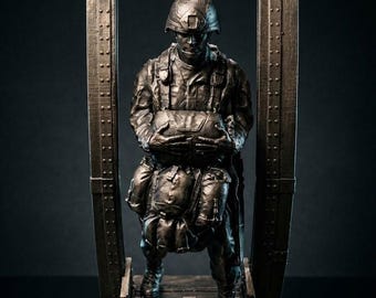 May include: A bronze-toned sculpture of a soldier in full combat gear, including a helmet and tactical vest, standing within a rectangular metal frame with visible rivets. The figure holds a large pack in front of the chest.