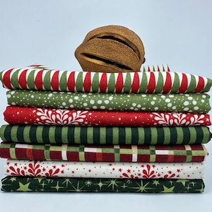 Könnte beinhalten: Ein Stapel weihnachtlicher Stoffquadrate in verschiedenen Mustern. Die Stoffe weisen rote, grüne und weiße Designs auf, darunter Streifen, Punkte, Schneeflocken und Sterne. Ein getrocknetes, braunes Dekorationselement liegt oben auf dem Stoffstapel.