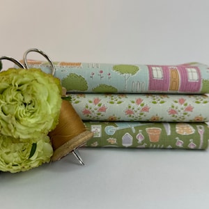 Könnte beinhalten: Ein Stapel von drei Stoffrollen mit Gartenmotiven. Die oberste Rolle zeigt ein Hausdesign, die mittlere Rolle florale Muster und die untere Rolle Gartengeräte. Eine Garnrolle und eine Schere befinden sich im Vordergrund.