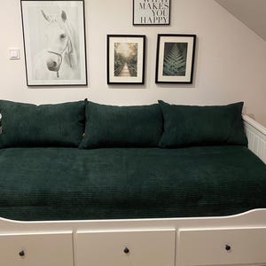 Może przedstawiać: Biała sofa z trzema poduszkami z ciemnozielonego sztruksu i pasującą narzutą. Na ścianie nad łóżkiem znajdują się oprawione obrazy, w tym portret konia i sceny przyrodnicze. Łóżko posiada trzy szuflady.