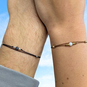 Puede incluir: Dos pulseras a juego con cordones negros y marrones, cada una con una sola cuenta. La pulsera de la izquierda tiene una cuenta gris oscura, mientras que la pulsera de la derecha tiene una cuenta blanca.
