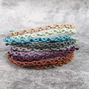May include: A stack of eight crocheted bracelets in various colours, including beige, teal, green, grey, purple, and brown. The bracelets are made with a simple, repeating pattern.