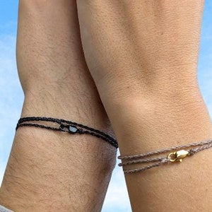 Peut inclure: Deux bracelets d'amitié assortis, l'un noir et l'autre gris, avec des fermoirs dorés. Les bracelets sont portés aux poignets de deux personnes dont les mains sont levées en l'air.