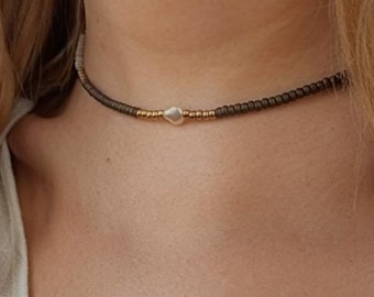 Può includere: Una collana girocollo composta da una serie di piccole perline grigio scuro, arricchita da una perla bianca irregolare centrale affiancata da due piccole sezioni di perline color oro metallizzato, indossata sopra un top di colore bianco.