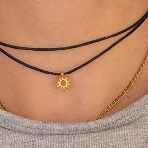 以下が含まれることがあります： 黒いコードチョーカーと金の太陽ペンダントが特徴のレイヤードネックレス。金のチェーンネックレスも見える。画像には「ARTWAVES design」の文字が入ったグレーのシャツが写っています。